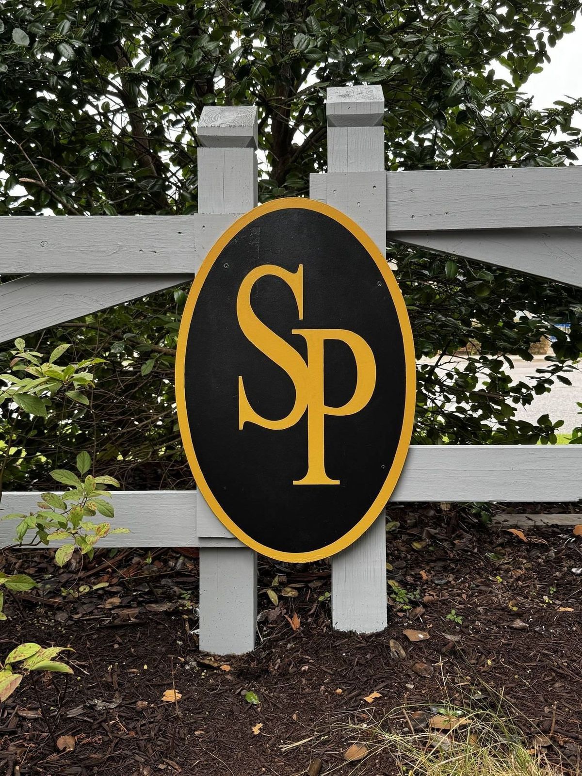 Oval neighborhood entrance sign with a gold monogram painted on black, set into a gray post and rail fence